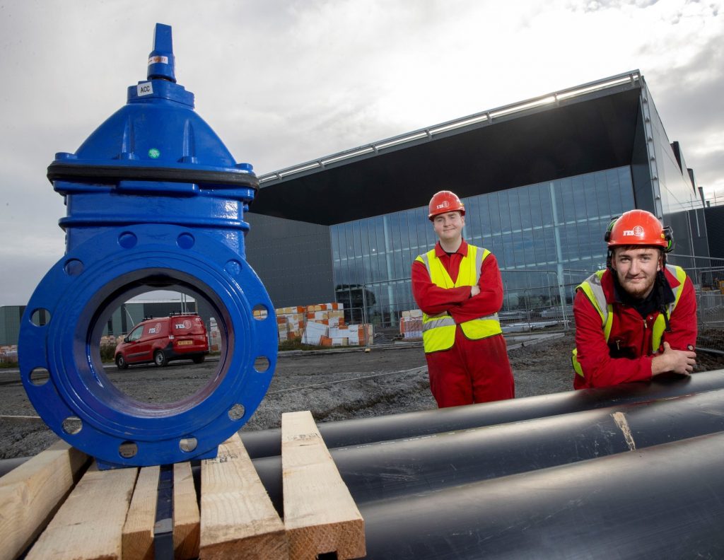 Two apprentices standing with parts for the low carbon heating network at AMIDS.
