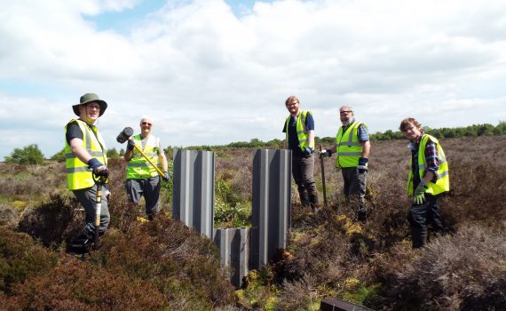 Decorative purposes. Photograph of five people in reflective jackets holding spades standing next to a new small metal dam, surrounded by peatland.