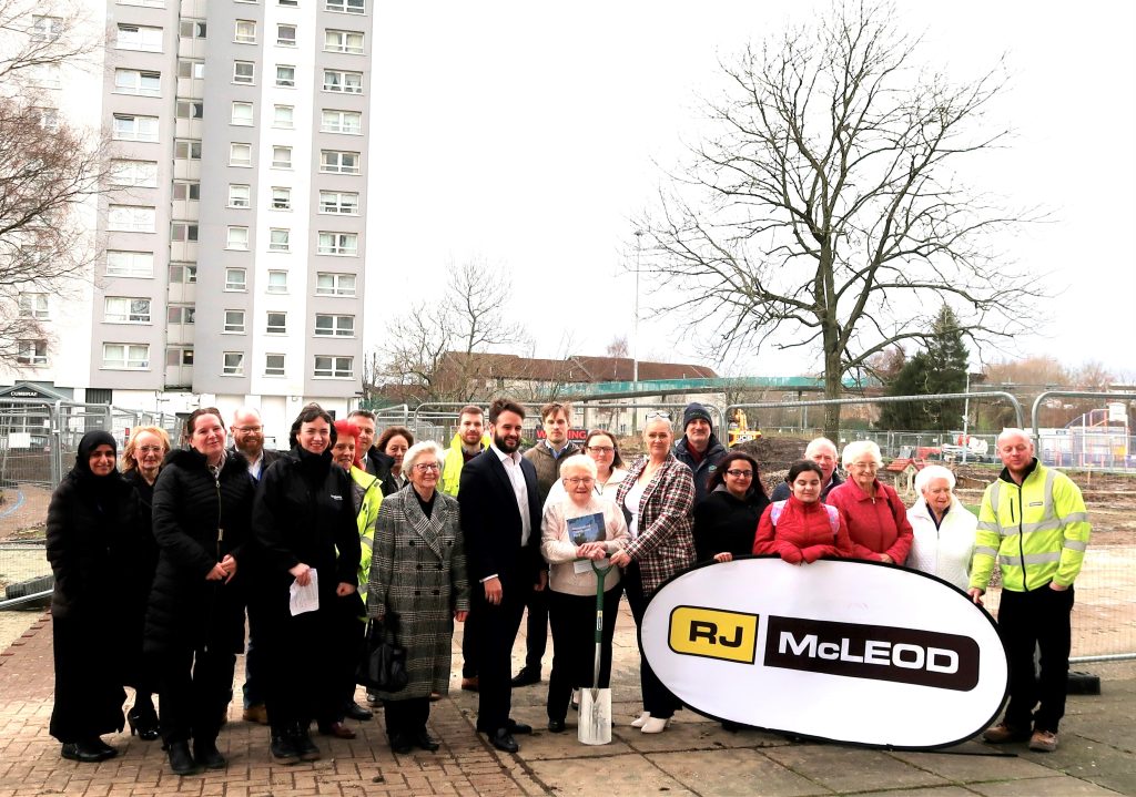 A group of around 25 funders and residents standing in front of fencing where the garden will be, to mark construction starting. The group are holding a sign which says "R.J. McLeod".