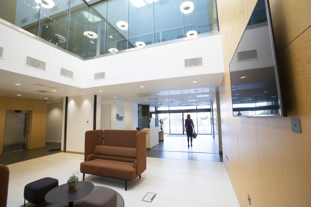 The inside atrium of The Greenlaw Works, looking out towards the main entrance door and angled upwards to show the atrium above. The image shows a modern-looking, bright workspace with clean, modern furniture and a television mounted on the wall of the ground floor.