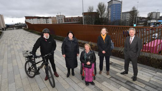 A photograph of 5 people, including Susan Aitken, standing on the new bridge. One person is holding a bicycle and another is in a wheelchair.