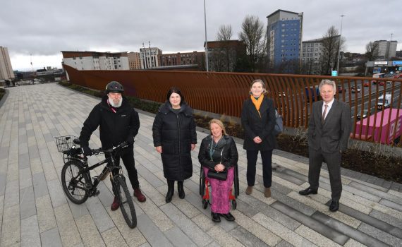 A photograph of 5 people, including Susan Aitken, standing on the new bridge. One person is holding a bicycle and another is in a wheelchair.
