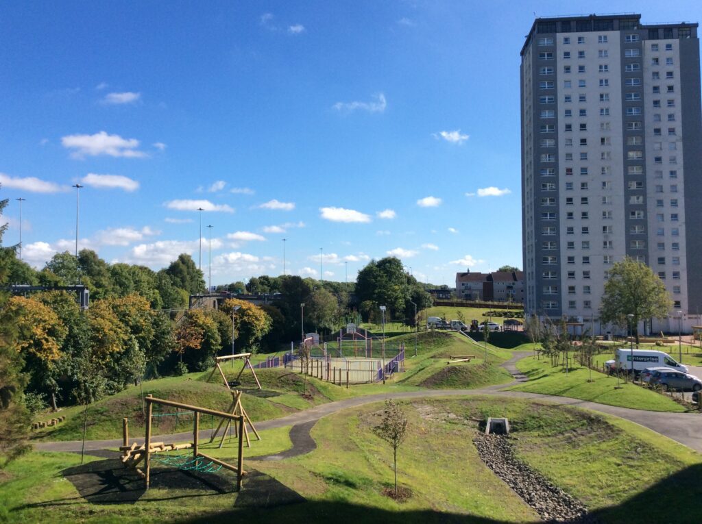 Looking down over the new gardens, showing the Queensland Court and Gardens high-rise flats in the back right. There is a children's play area in the bottom left, a basketball court in the centre of the gardens, and a winding path through the new greenspace.