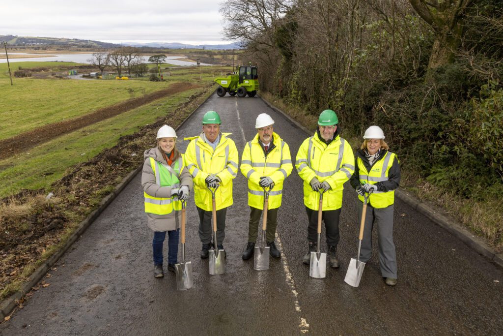 Five people in reflective jackets, hard hats and safety gear stand on Aurs Road holding shovels, at the front of the picture. Aurs Road stretches out behind them. Photograph depicts: Cllr Owen O'Donnell, Leader, East Renfrewshire Council; Caitriona McAuley, Director of Environment, East Renfrewshire Council; Leo Martin, Managing Director of Civil Engineering, GRAHAM; Jim Armour, Contracts Manager, GRAHAM; Karen McGregor, Scotland Director, Sustrans