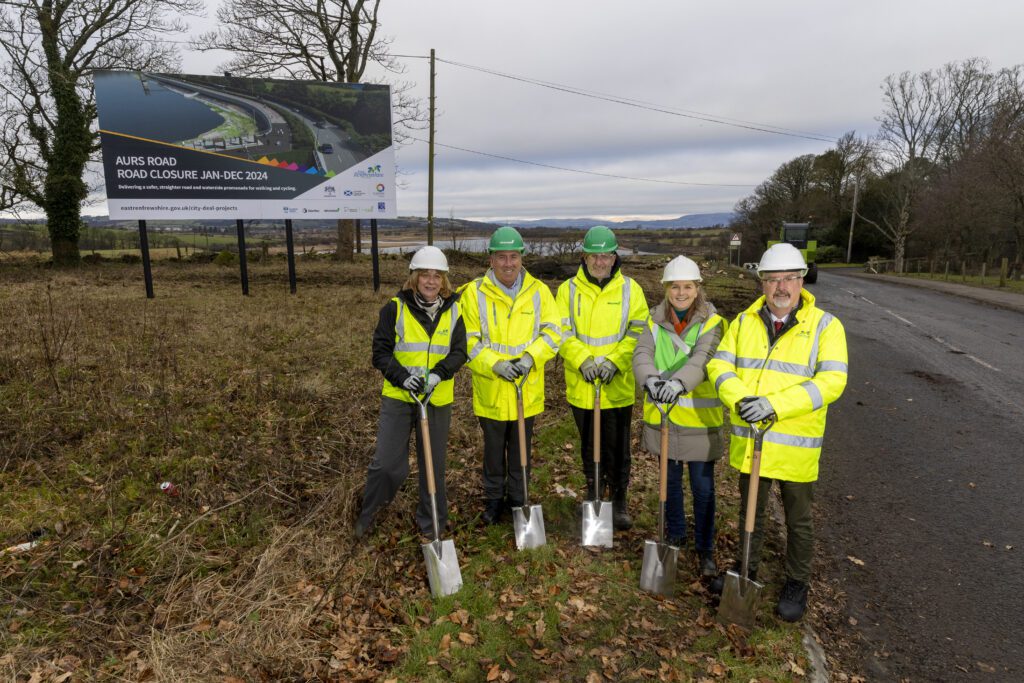 Five people in reflective jackets, hard hats and safety gear stand on Aurs Road holding shovels, in front of "Aurs Road - Road Closure" signage. Photograph depicts: Cllr Owen O'Donnell, Leader, East Renfrewshire Council; Caitriona McAuley, Director of Environment, East Renfrewshire Council; Leo Martin, Managing Director of Civil Engineering, GRAHAM; Jim Armour, Contracts Manager, GRAHAM; Karen McGregor, Scotland Director, Sustrans
