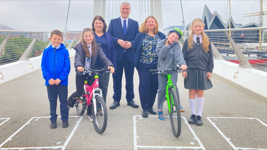 Four schoolchildren, including two on bikes, standing on the new Govan to Partick bridge in front of: (from left to right) Councillor Susan Aitken, Tom Arthur, Kirsty McNeil