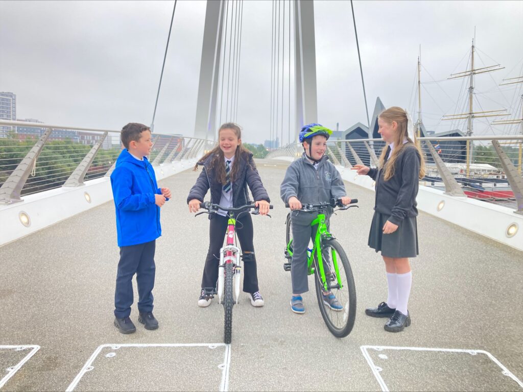 Four schoolchilren standing on the new Govan to Partick bridge, two on bikes. They are speaking to each other and smiling/laughing. The Riverside Museum and Tall Ship are visible in the back right of the photo, behind the bridge.